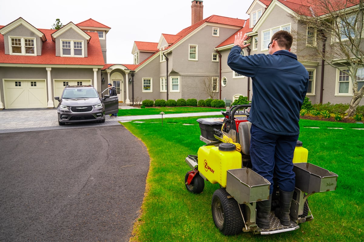 lawn care technician waves to homeowner while fertilizing grass